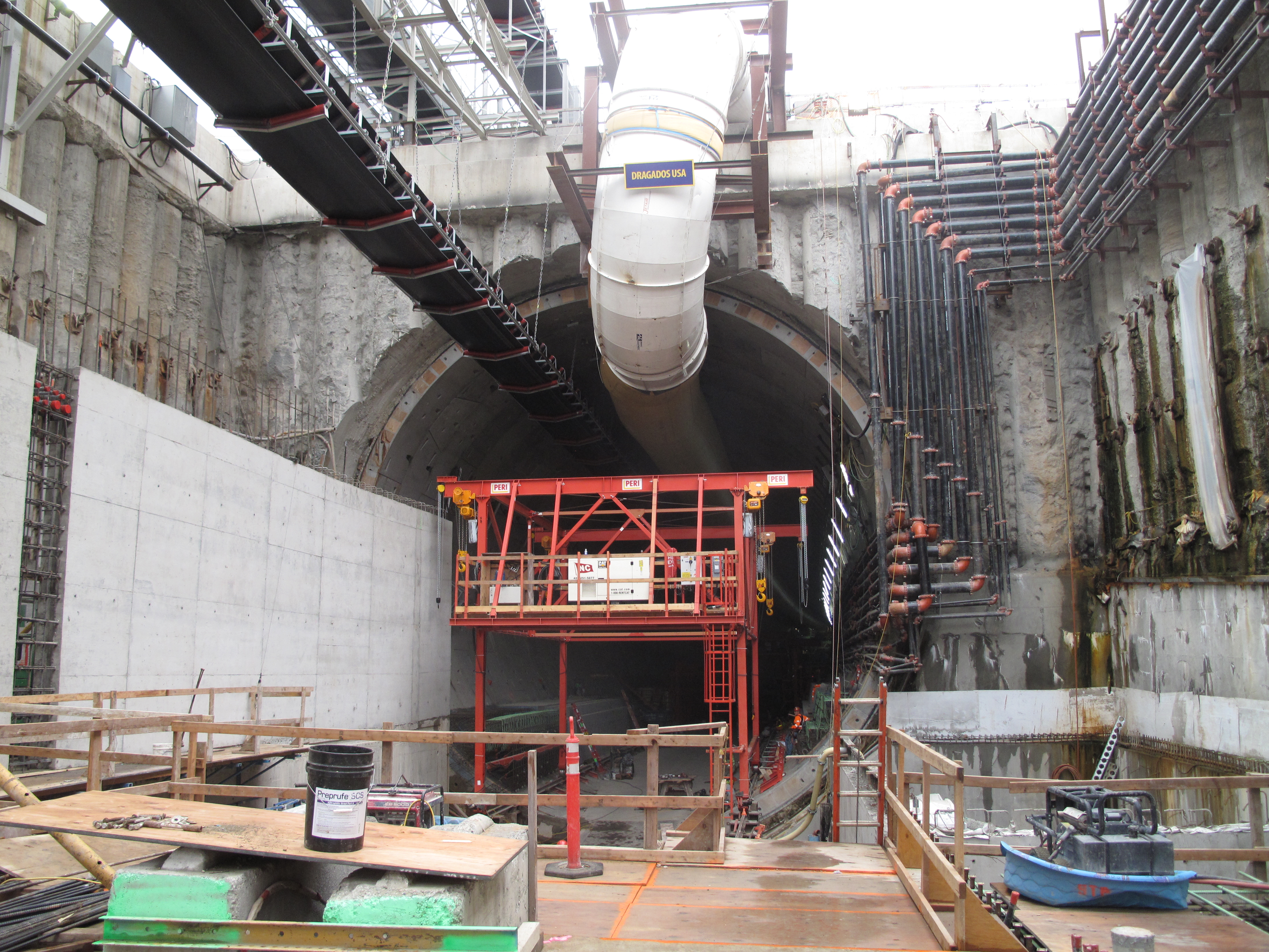 caption: The south tunnel entrance has been lined with concrete. Along the top of the tunnel, on the left a black conveyer belt hauls out excavated soil, while the white pipe funnels in air (or sucks it out in the case of a fire).