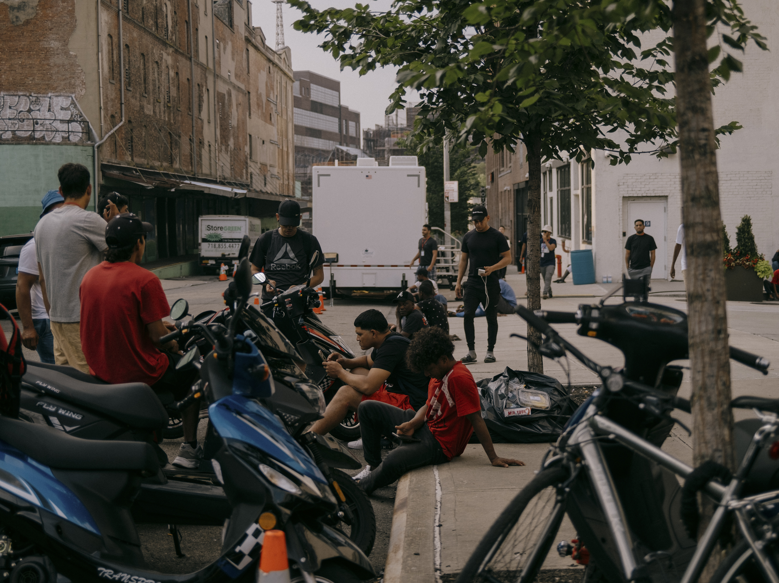 caption: Migrants at the Clinton Hill Shelter seek relief from the overcrowded confinements of 47 Hall Street. Residents working as delivery drivers wait for orders to come in, Brooklyn, N.Y., on July 19, 2023.