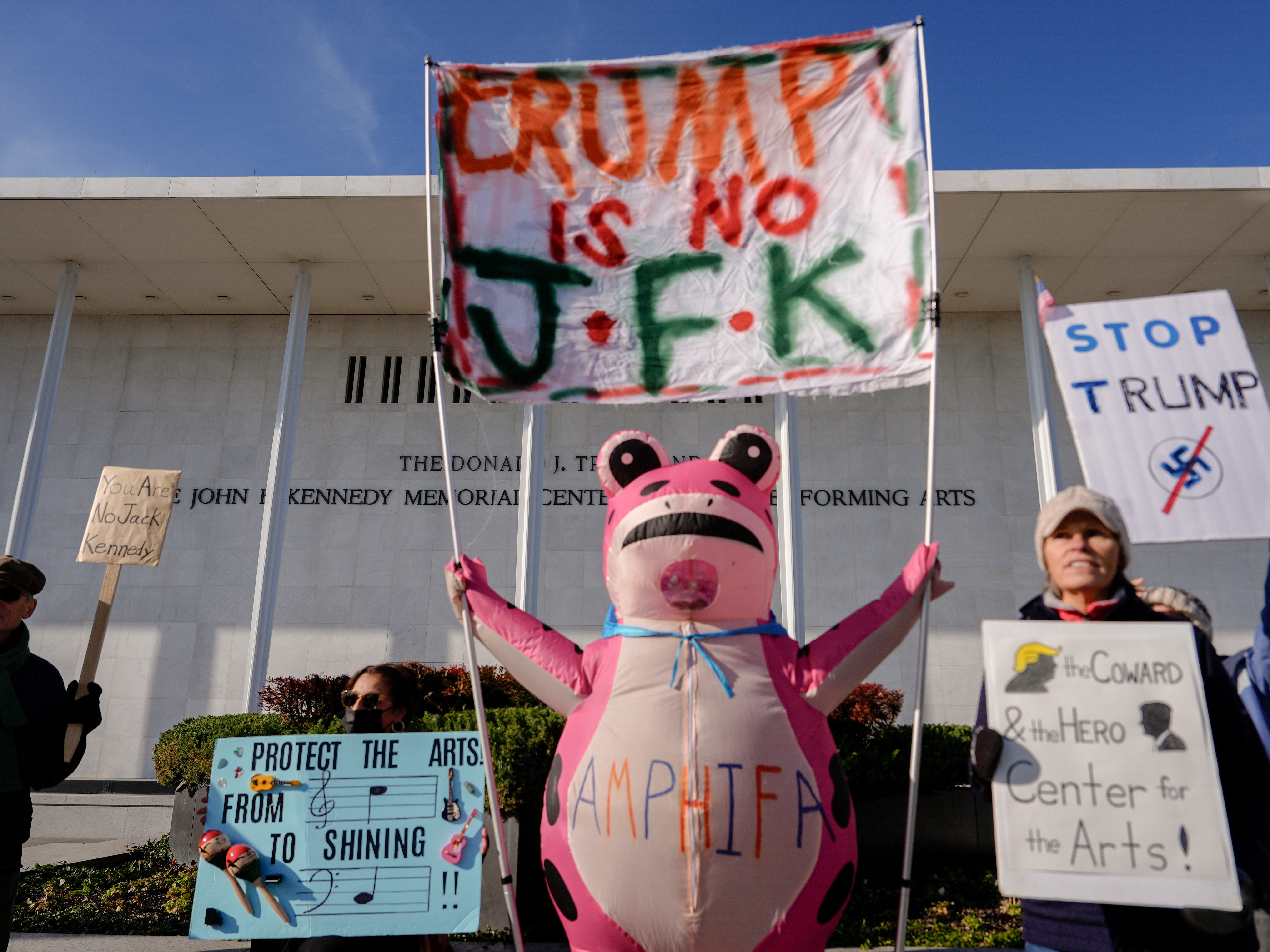 caption: Demonstrators, including Nadine Siler, of Waldorf, Md., dressed in a pink frog costume, hold up signs at a designated protest point in front of the John F. Kennedy Memorial Center for the Performing Arts, a day after a Trump-appointed board voted to add President Donald Trump's name to the Kennedy Center, Saturday, Dec. 20, 2025, in Washington. (AP Photo/Julia Demaree Nikhinson)