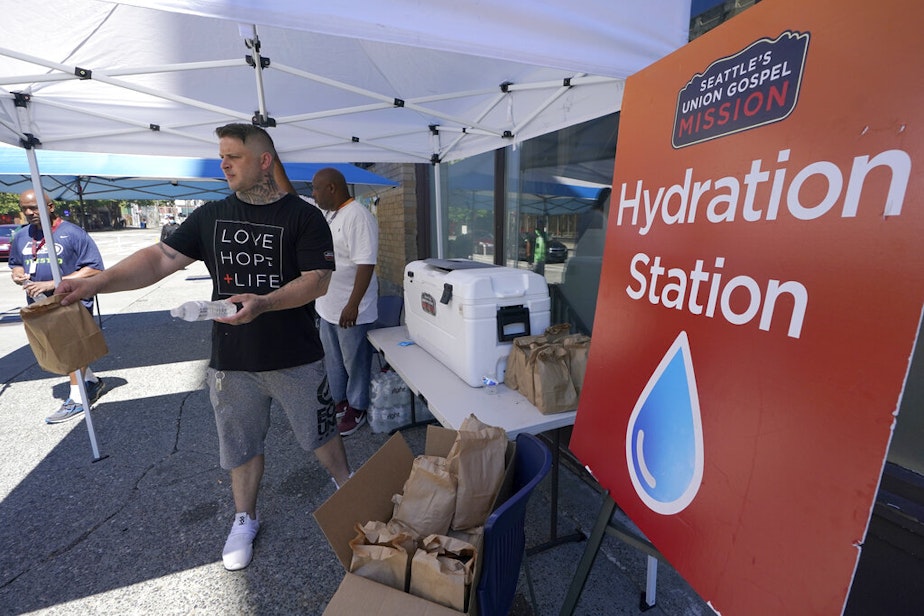 caption: Carlos Ramos hands out bottles of water and sack lunches, Monday, June 28, 2021, as he works at a hydration station in front of the Union Gospel Mission in Seattle.