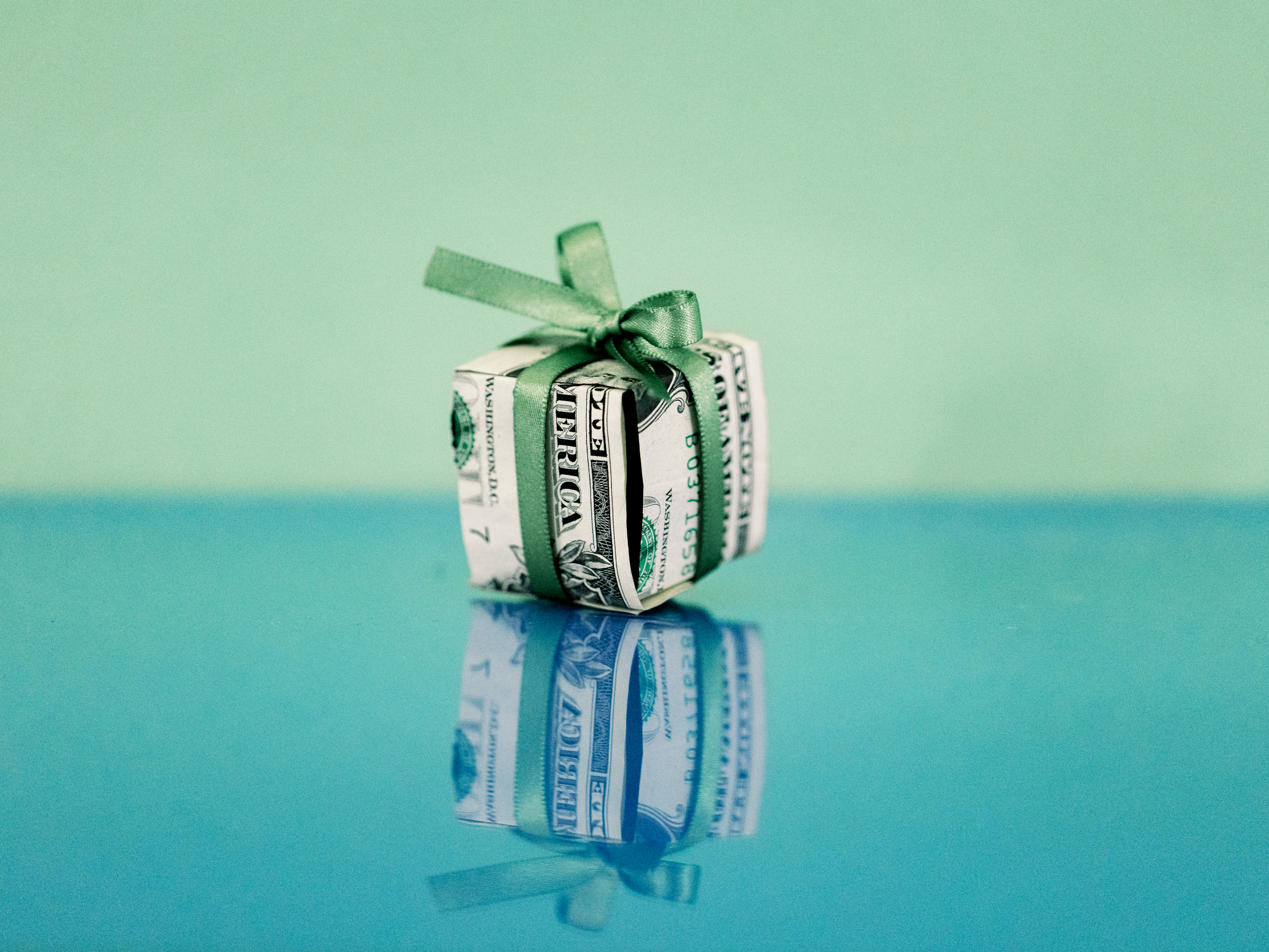 A U.S. dollar bill is folded into the shape of a gift box and tied with a green ribbon. The gift box is photographed against a blue and green background and the box is reflected in the surface below.