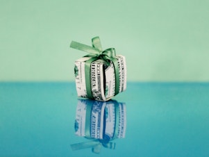 A U.S. dollar bill is folded into the shape of a gift box and tied with a green ribbon. The gift box is photographed against a blue and green background and the box is reflected in the surface below.