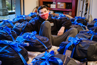 caption: Harry McGovern sitting in the garage of one of his wish granters, Lynne Witham, surrounded by the first 25 packages created by The First Night Project in October 2020.