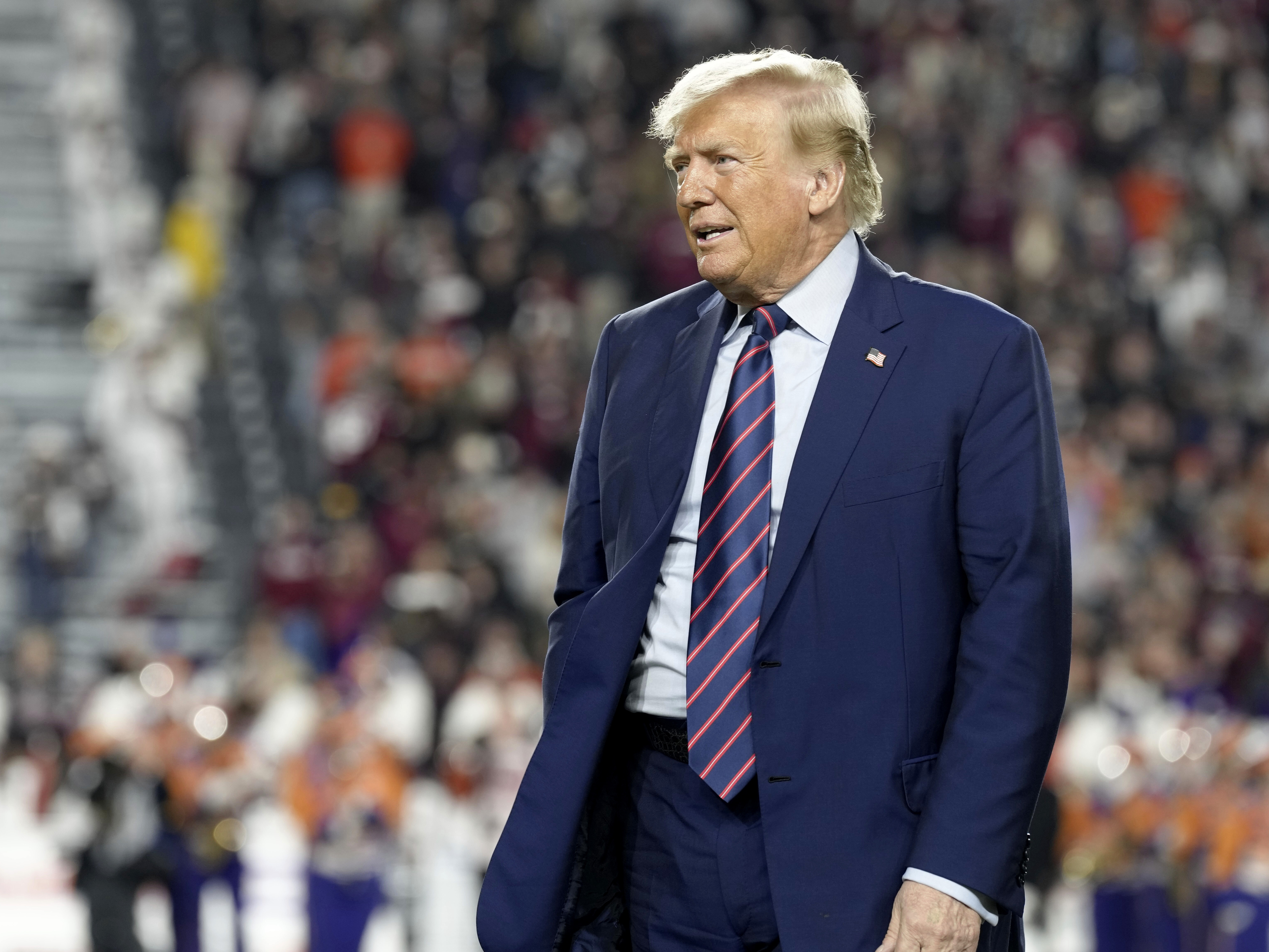 caption: Republican presidential candidate and former President Donald Trump stands on the field during halftime in an NCAA college football game between the University of South Carolina and Clemson Saturday, Nov. 25, 2023, in Columbia, S.C.