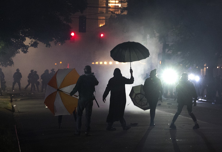 caption: A group of protesters stand in front of hundreds of other protesters as Seattle police officers dressed in riot gear shine lights following the use tear gas, pepper spray and flash-bang grenades on the fifth day of protests following the violent police killing of George Floyd on Tuesday, June 2, 2020, at the intersection 11th and Pine Streets in Seattle.