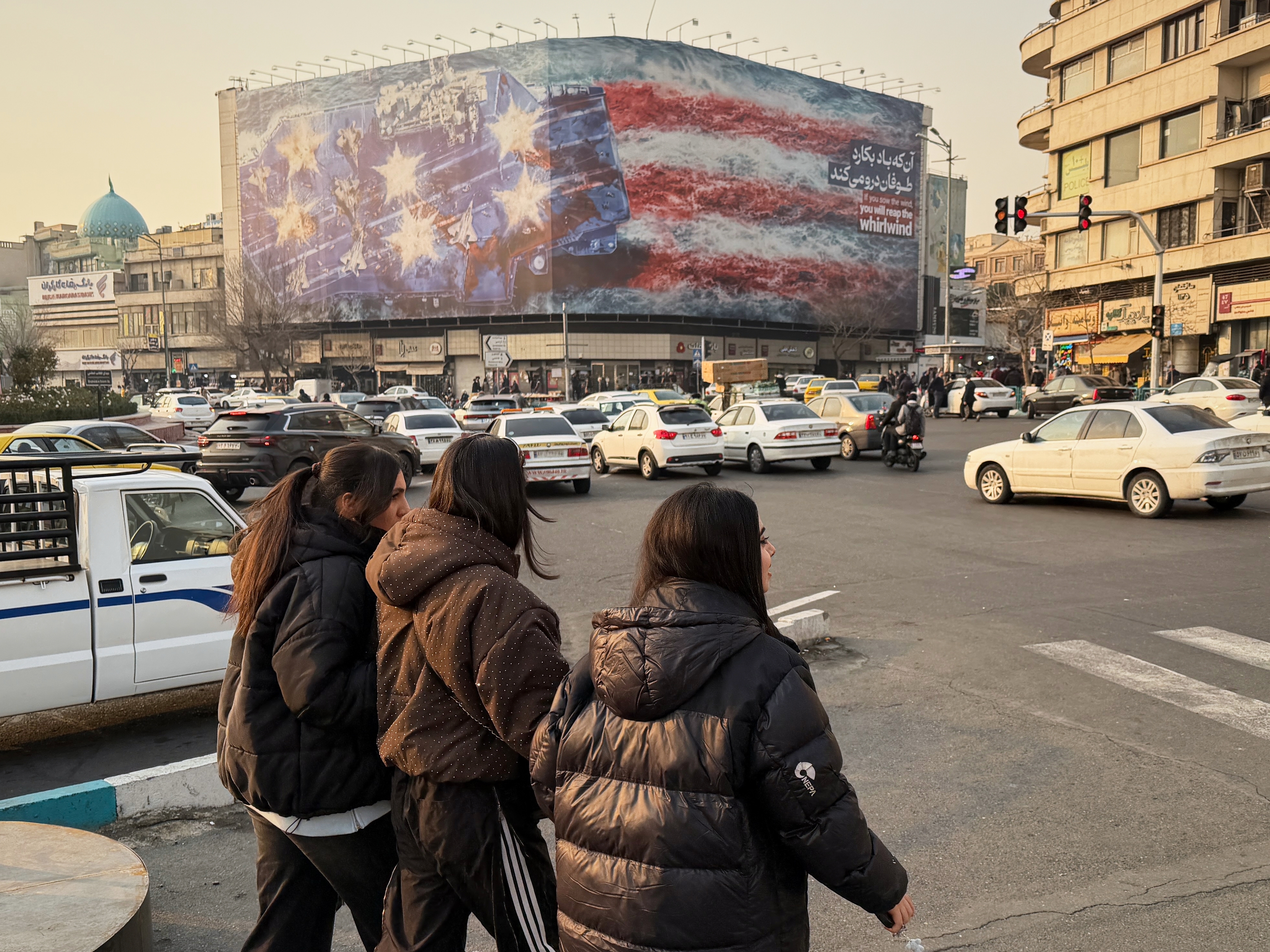 caption: People walk in front a billboard with graphic showing a U.S aircraft carrier with damaged fighter jets on its deck, and sign reading in Farsi and English: "If you sow the wind, you'll reap whirlwind," at the Enqelab-e-Eslami (Islamic Revolution) square, in Tehran, Iran, Sunday, Jan. 25, 2026.