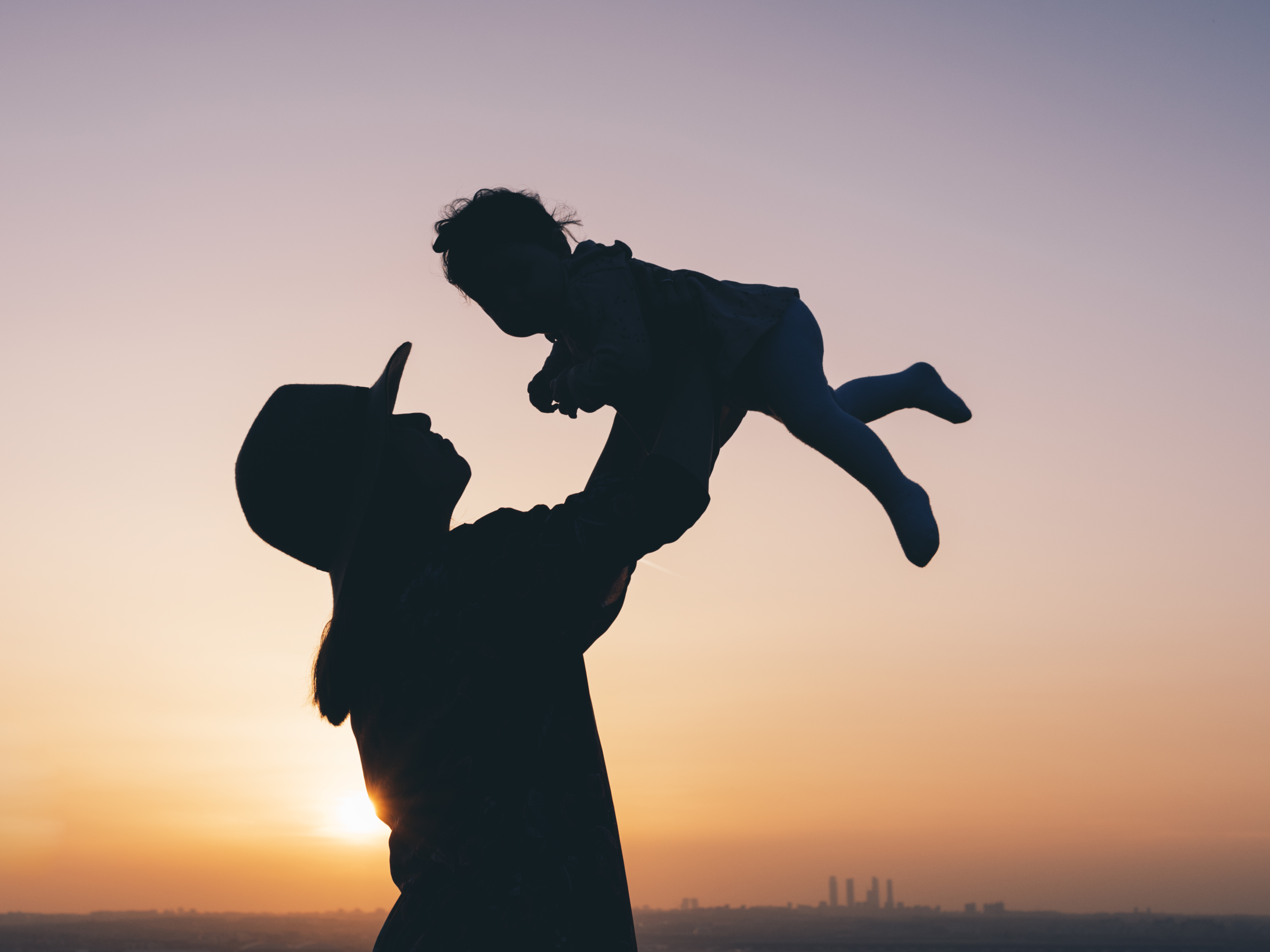 Silhouette of a mother lifting her baby in the air outdoors during a beautiful sunset. She is wearing a hat.