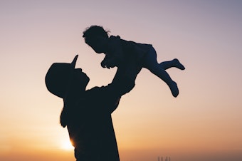 Silhouette of a mother lifting her baby in the air outdoors during a beautiful sunset. She is wearing a hat.