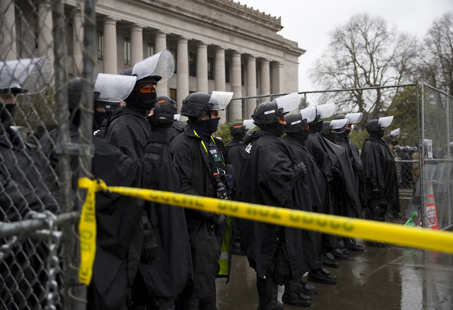 caption: Police guard the Washington State Capitol building on Monday, January 11, 2021, in Olympia. Armed protesters gathered on the opposite side of the fence on the first day of the legislative session in Olympia. 