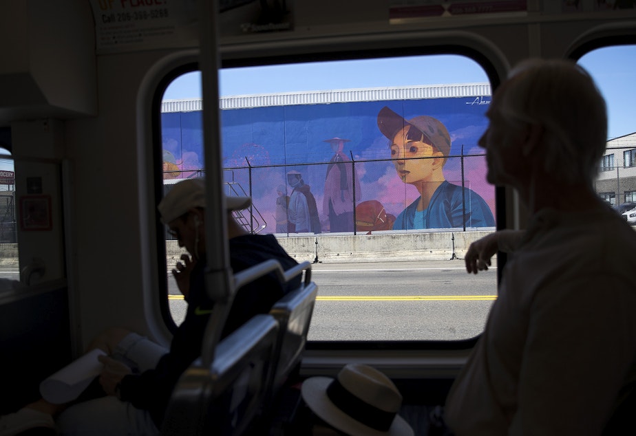 caption: A mural painted by artist Andrew Hem is shown through the window of the light rail as commuters ride by on Tuesday, August 15, 2017, along the SODO Track in Seattle. 