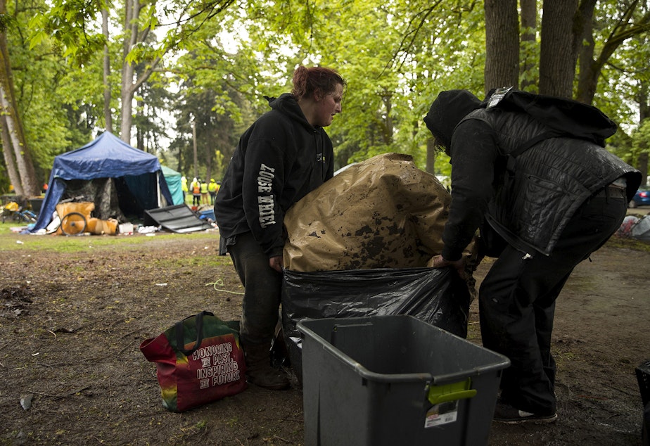 caption: Kelsey, left, and Tyler, right, work to secure tarps into a garbage bag as the city of Seattle removed unhoused people and their belongings from an encampment at Woodland Park on Tuesday, May 10, 2022, in Seattle. 