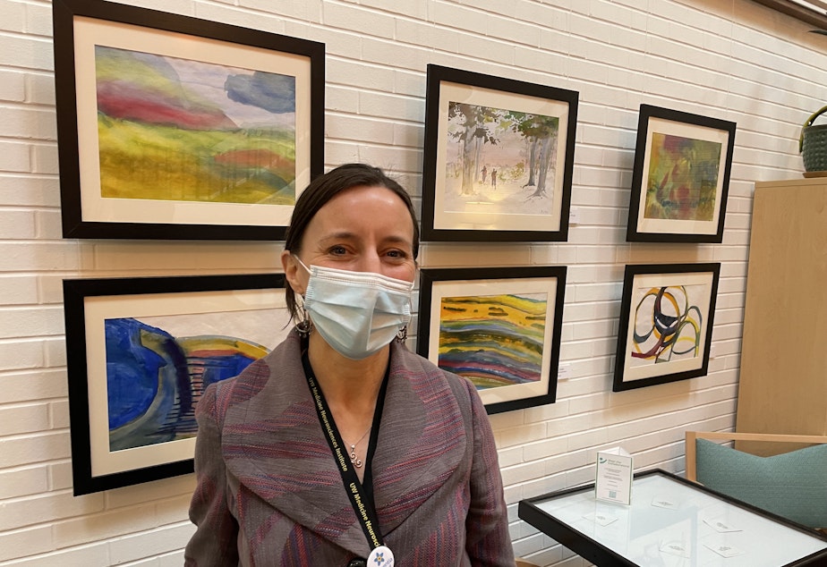 caption: Director of the Memory Hub, Marygrace Becker, stands in front of a wall of watercolor paintings 