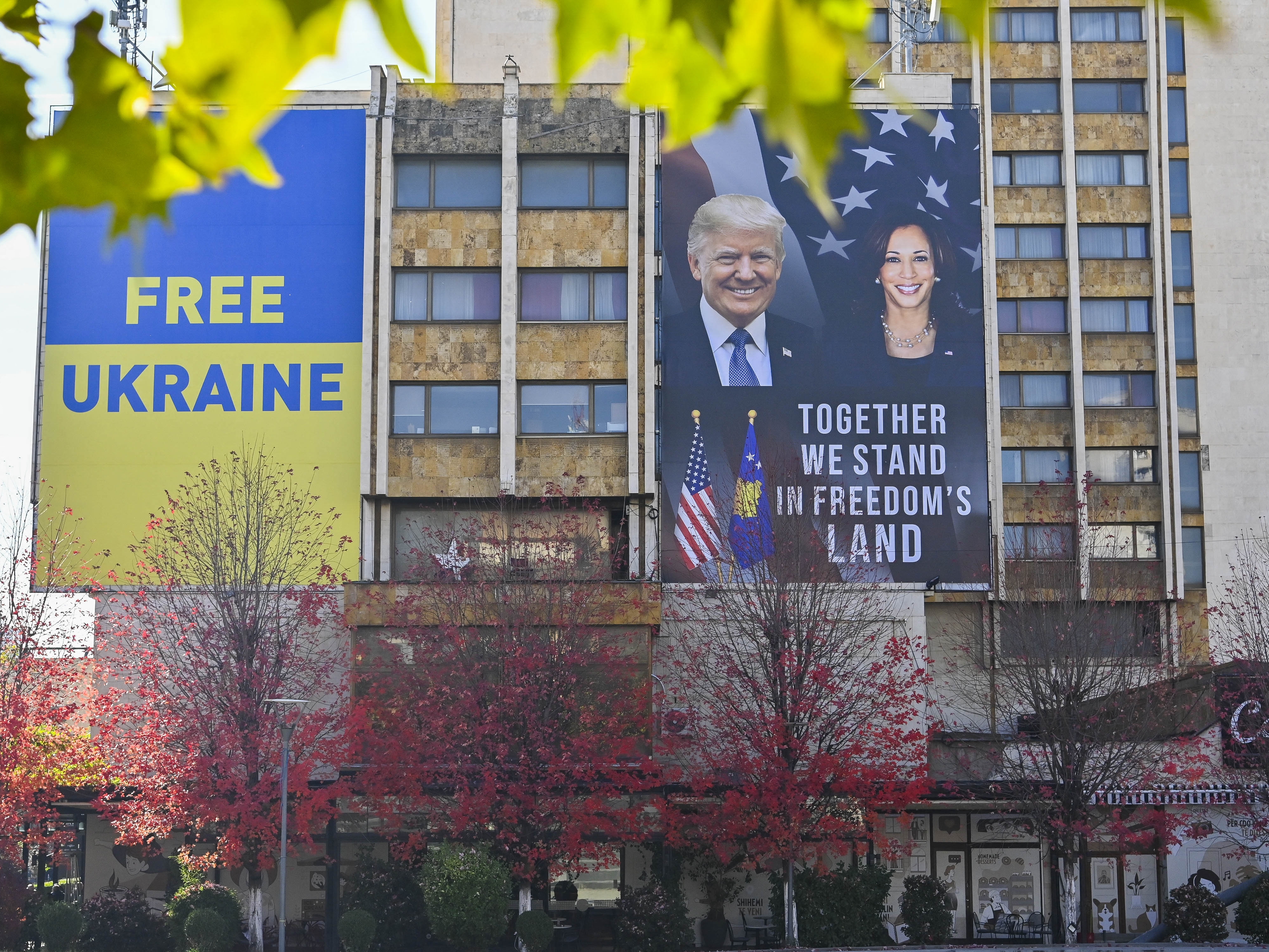 caption: A large banner bearing the portrait of US Vice President and Democratic presidential candidate Kamala Harris and former US President and Republican presidential candidate Donald Trump is displayed next to a banner reading "Free Ukraine" on the facade of a hotel in Pristina on November 6 , 2024.