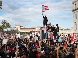 caption: Bad Bunny (holding flag), singer Ricky Martin (in black hat) and Residente (in blue hat) join demonstrators in protest against Puerto Rico Gov. Ricardo Roselló in Old San Juan.