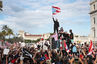 caption: Bad Bunny (holding flag), singer Ricky Martin (in black hat) and Residente (in blue hat) join demonstrators in protest against Puerto Rico Gov. Ricardo Roselló in Old San Juan.