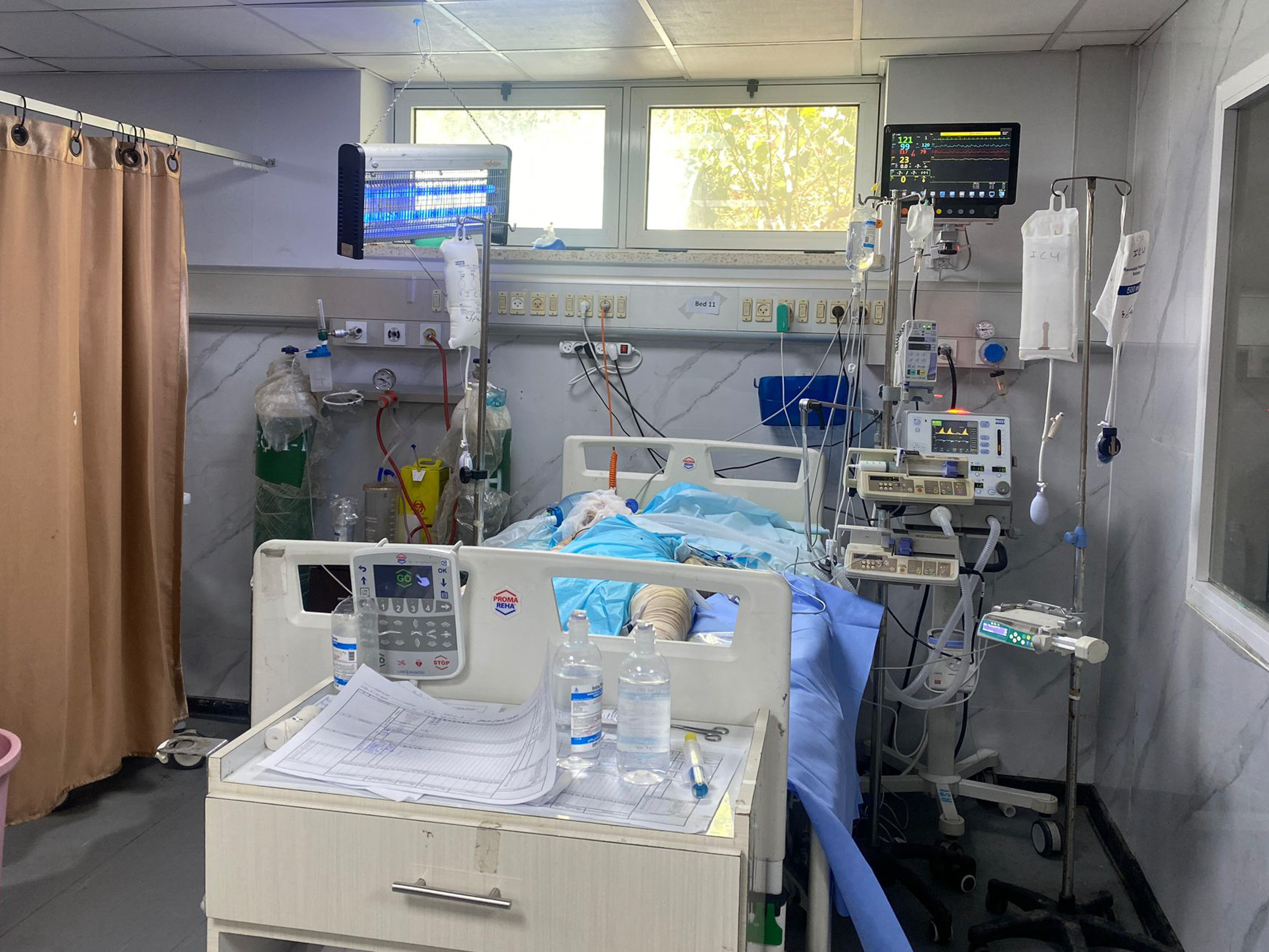 caption: A patient in European Gaza hospital this weekend — the last functioning hospital in Rafah. Staff placed an insect zapper above his bed to keep away the flies.