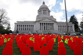 caption: Red mock tombstones represent the more than 1,000 people who took their lives by suicide in Washington state in 2017, displayed Tuesday, March 12, 2019 at the Capitol in Olympia. The yellow vests on some of the markers indicate the victim was from the construction industry, and similar markers were used to designate youth and military veterans. 