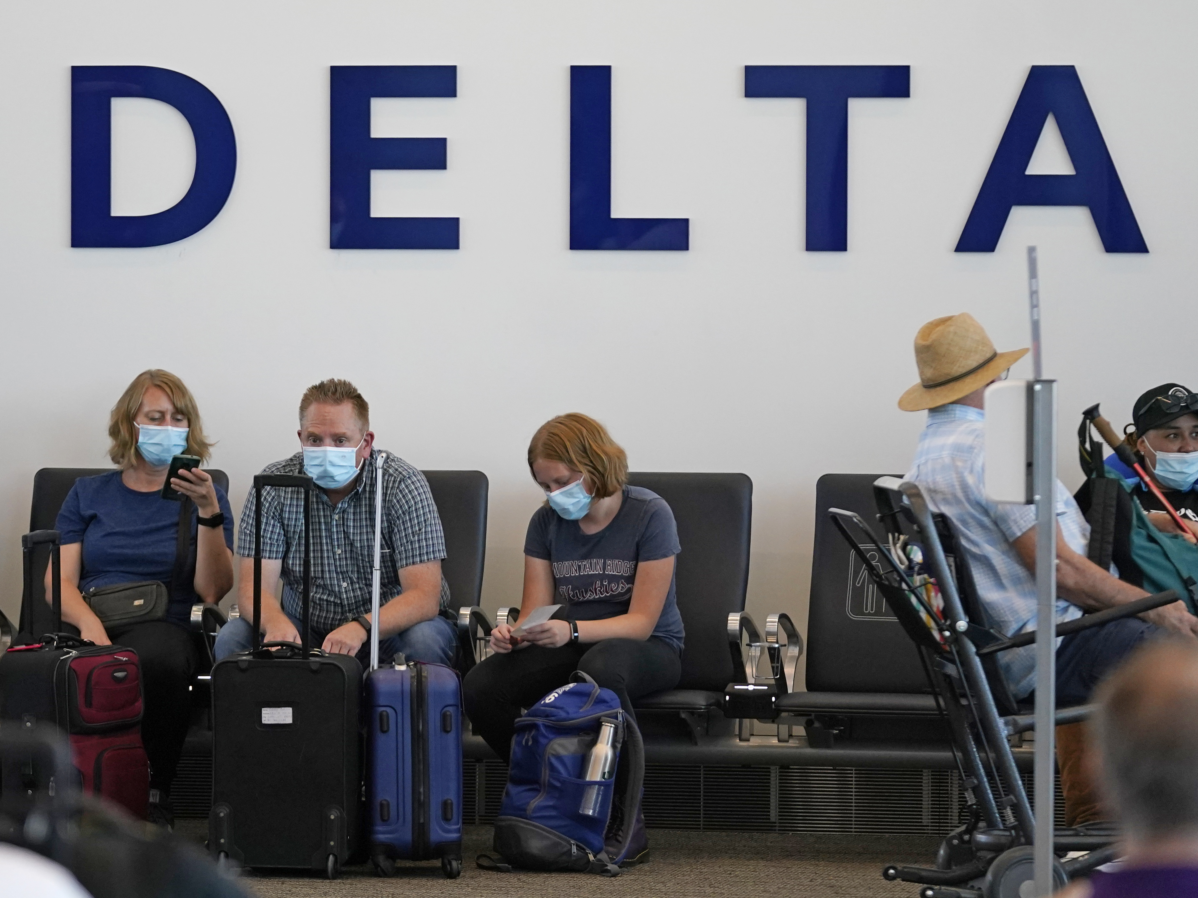 caption: People sit under Delta sign at Salt Lake City International Airport on July 1, 2021. Delta Air Lines won't force employees to get vaccinated, but it's going to make unvaccinated workers pay a $200 monthly charge.