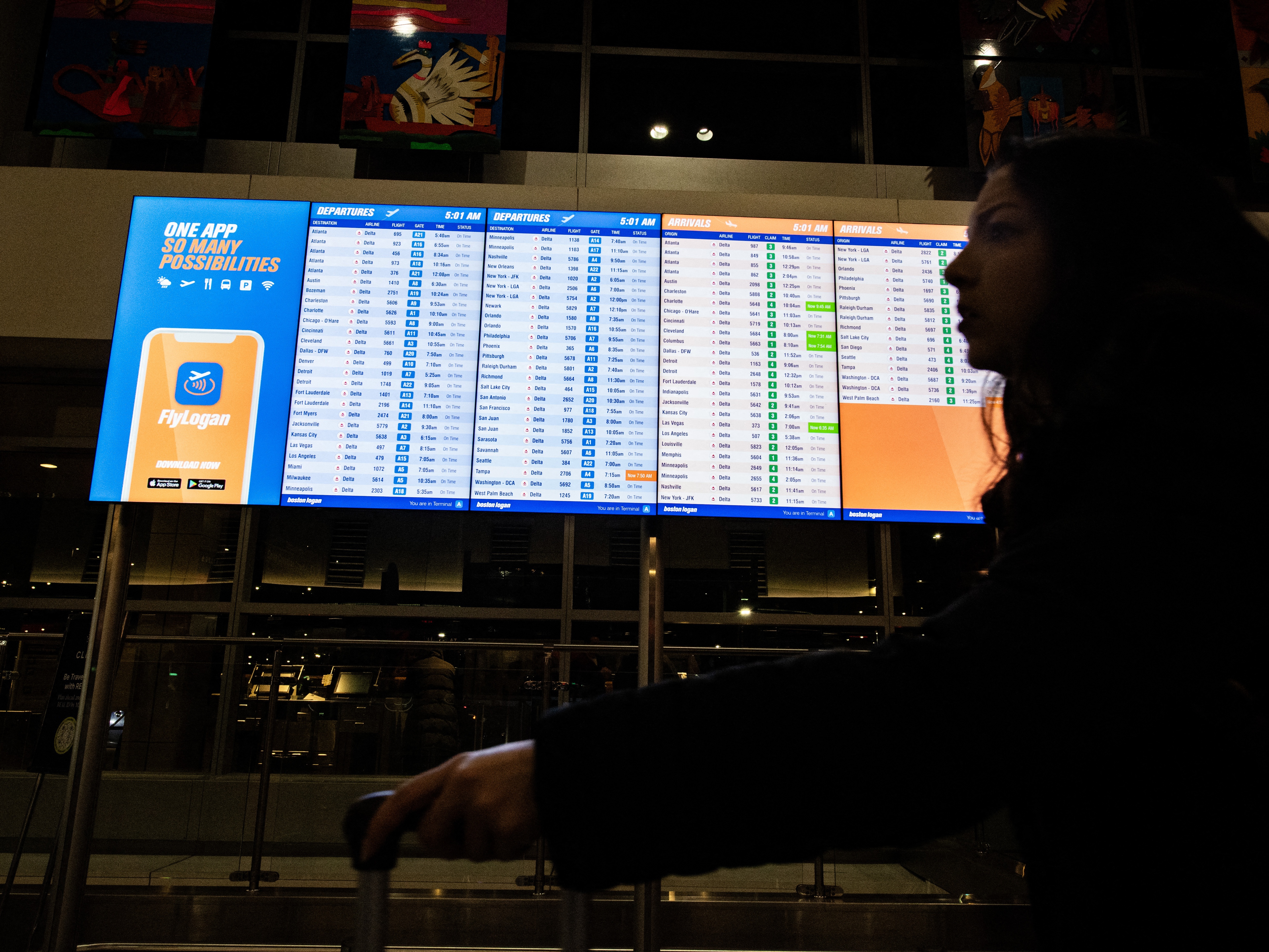 caption: A person walks by an arrival and departure board at Boston's Logan Airport on Dec. 23, 2024.