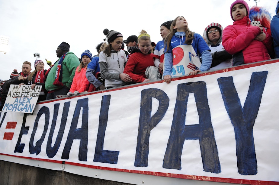 caption: Fans stand behind a large sign for equal pay for the women's soccer team during an international friendly soccer match in 2016. (Jessica Hill/AP)