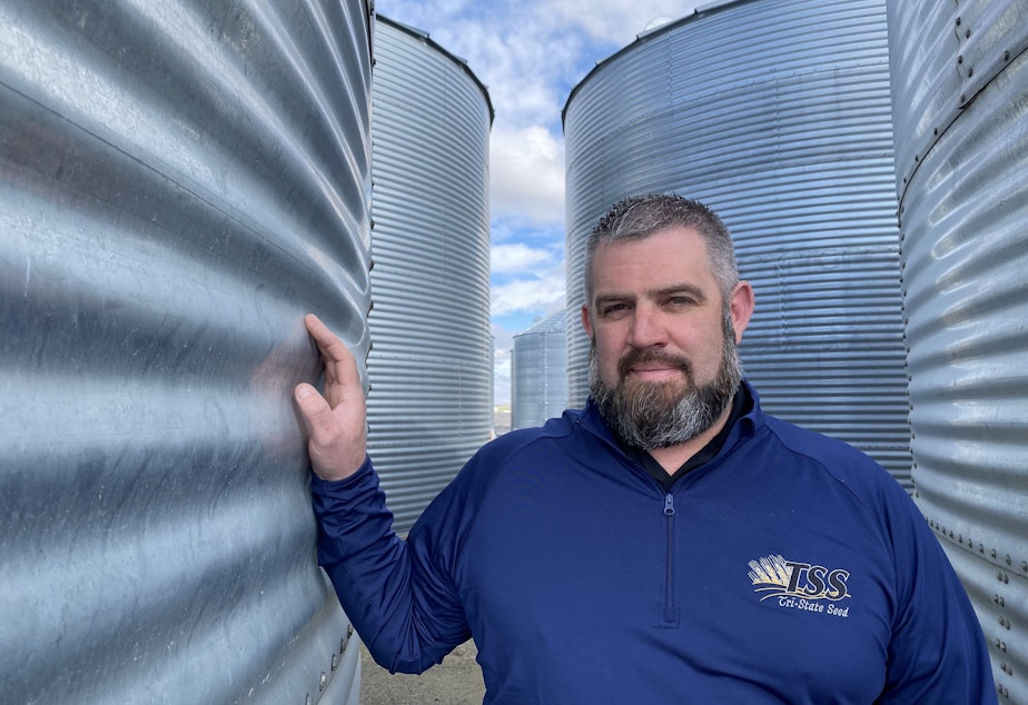caption: Mason Douglass leans on one of his thirty seed bins at Tri-State Seed outside of Connell, Wash.