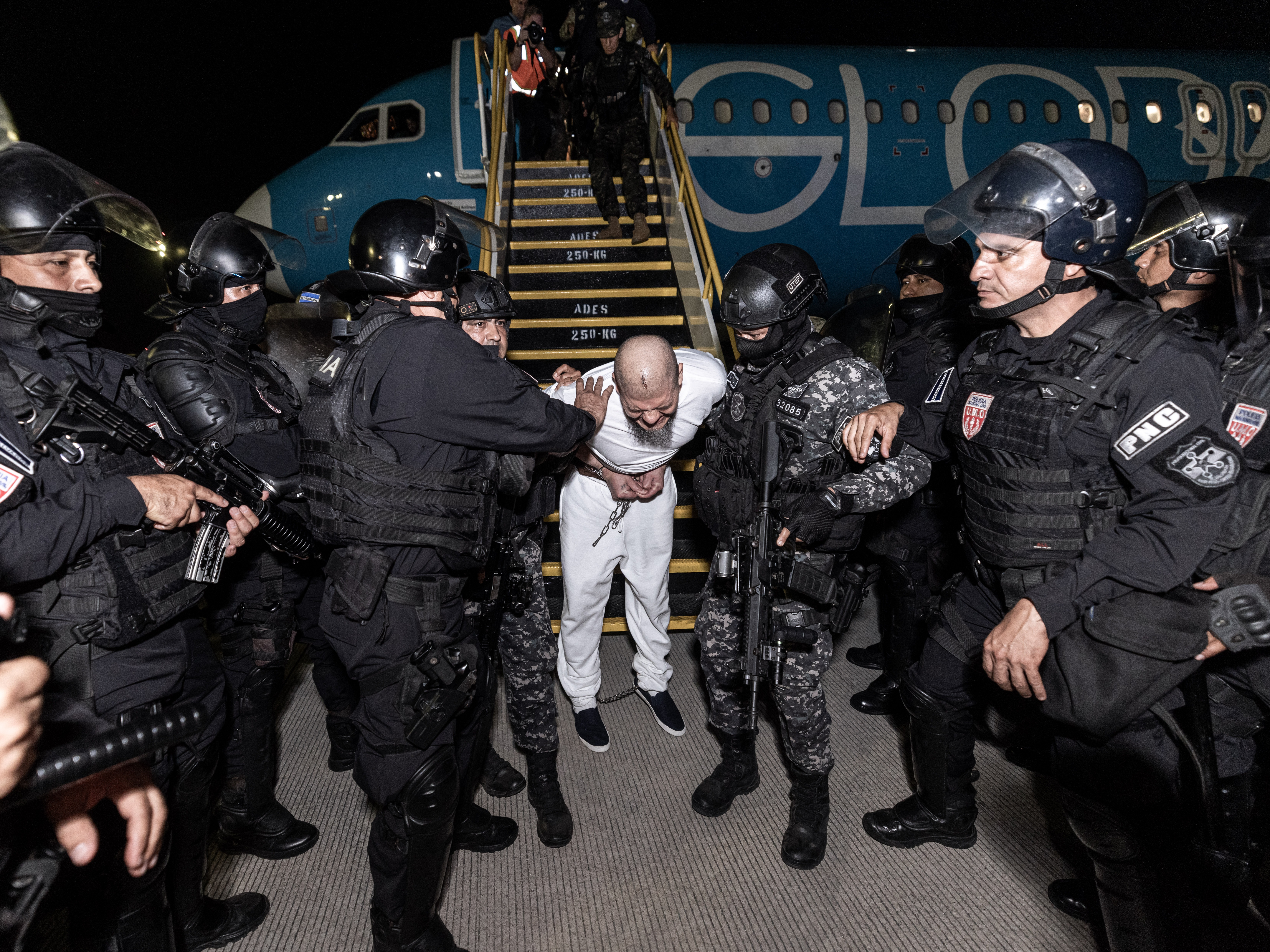 caption: Guards escort one of the hundreds of alleged members of the 'Tren De Aragua' and Mara Salvatrucha gangs who were deported from the U.S. to El Salvador.