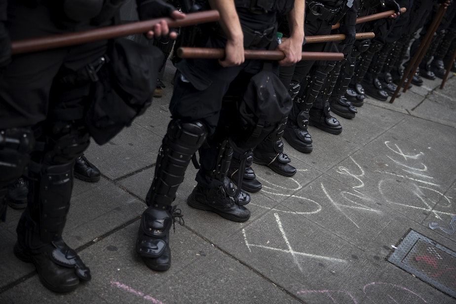 caption: Seattle police officers hold their batons while standing in a police line next to chalk writing on the sidewalk that says 'fuck the police,' on the fifth day of protests following the violent police killing of George Floyd on Tuesday, June 2, 2020, at the intersection 11th and Pine Streets in Seattle. 