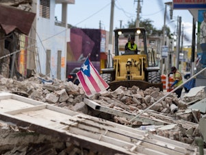 caption: A Puerto Rican flag waves on top of a pile of rubble as debris is removed from a main road in Guánica.