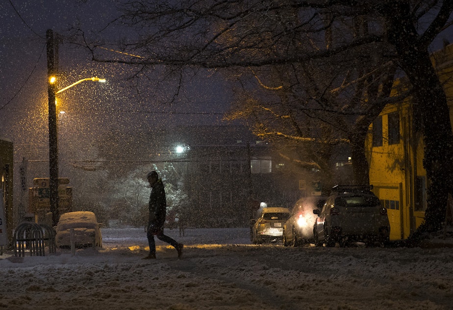 caption: A pedestrian crosses Ballard Avenue Northwest as snow falls on Monday, February 11, 2019, in Seattle.