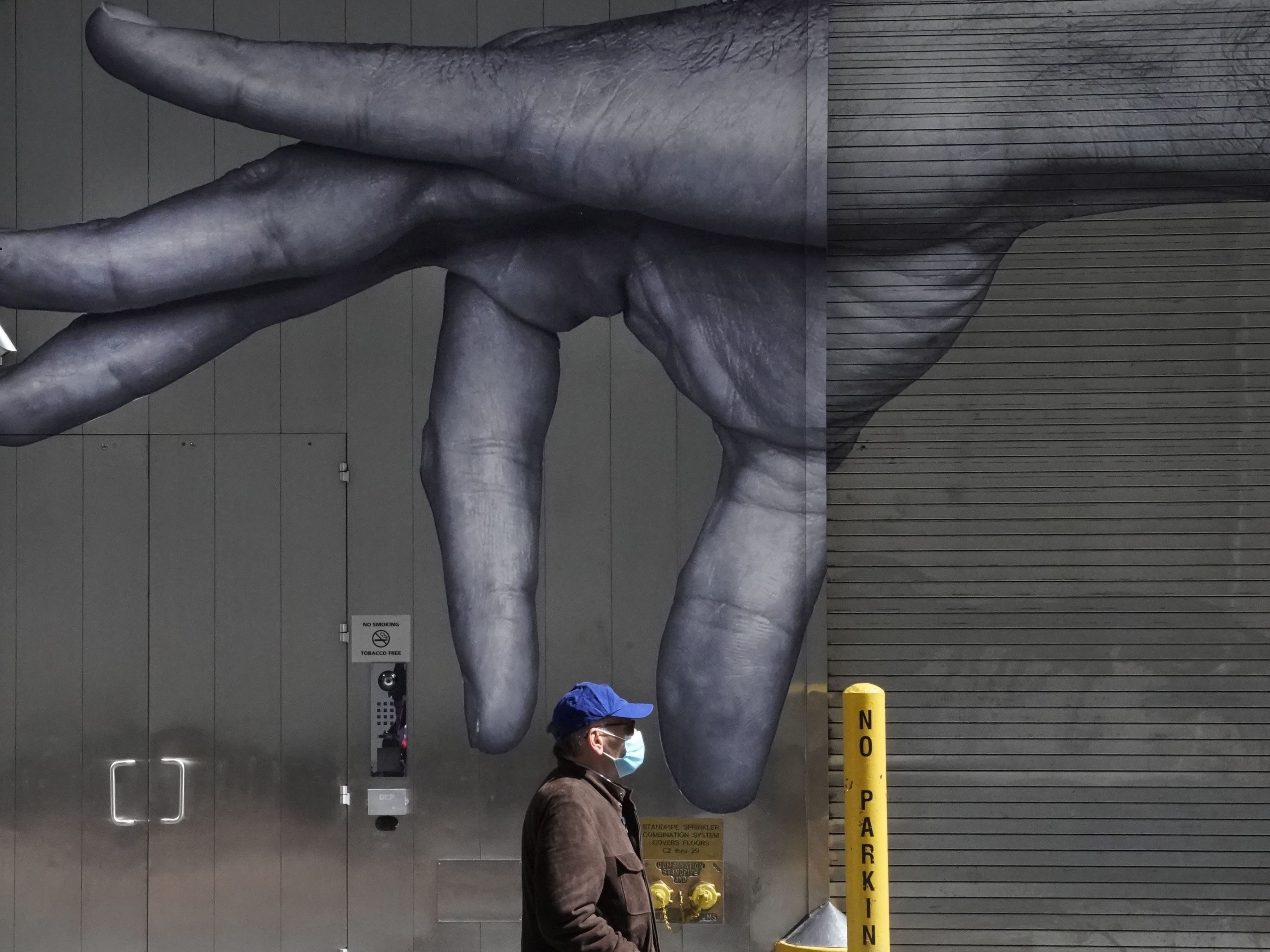 caption: A man in a mask walks past a mural on the side of a building in New York City in April.