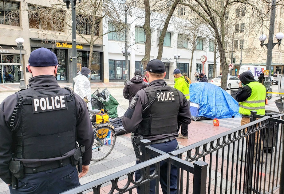 caption: Seattle Police officers watch Parks Department crews remove tents from Westlake Park on Friday, March 11, 2022.