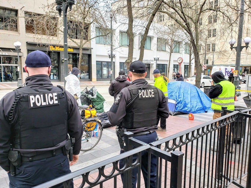 caption: Seattle Police officers watch Parks Department crews remove tents from Westlake Park on Friday, March 11, 2022.