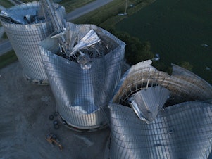 caption: Damaged grain bins are shown at the Heartland Co-Op grain elevator on Aug. 11, 2020, in Malcom, Iowa. Some people are still recovering a year after the 2020 derecho caused $11 billion in damage across the Midwest.