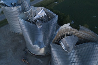 caption: Damaged grain bins are shown at the Heartland Co-Op grain elevator on Aug. 11, 2020, in Malcom, Iowa. Some people are still recovering a year after the 2020 derecho caused $11 billion in damage across the Midwest.