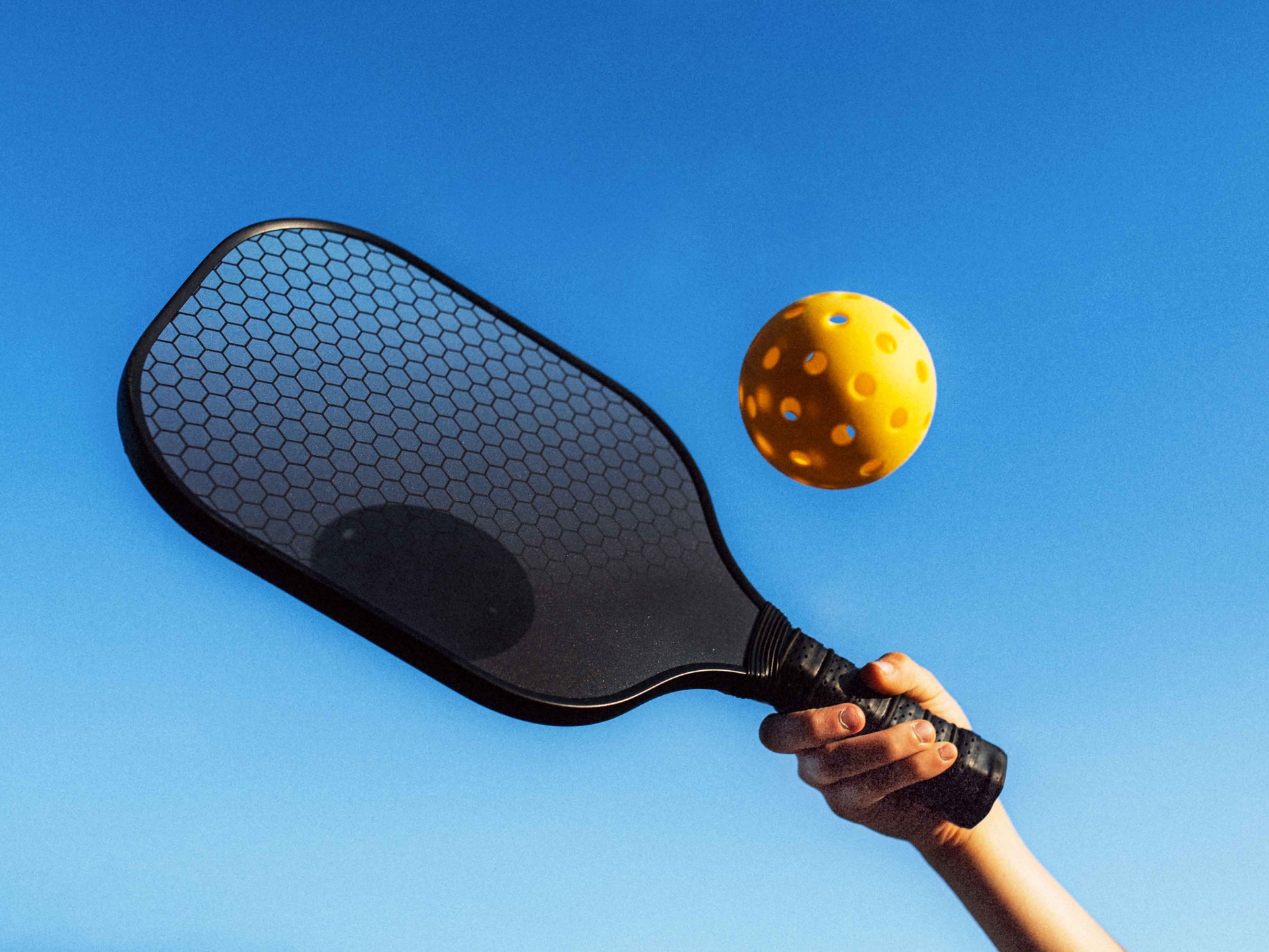 caption: A hand holds a paddle against a clear blue sky, preparing to hit a yellow pickleball. The scene suggests a bright and sunny day, with the pickleball clearly in motion.