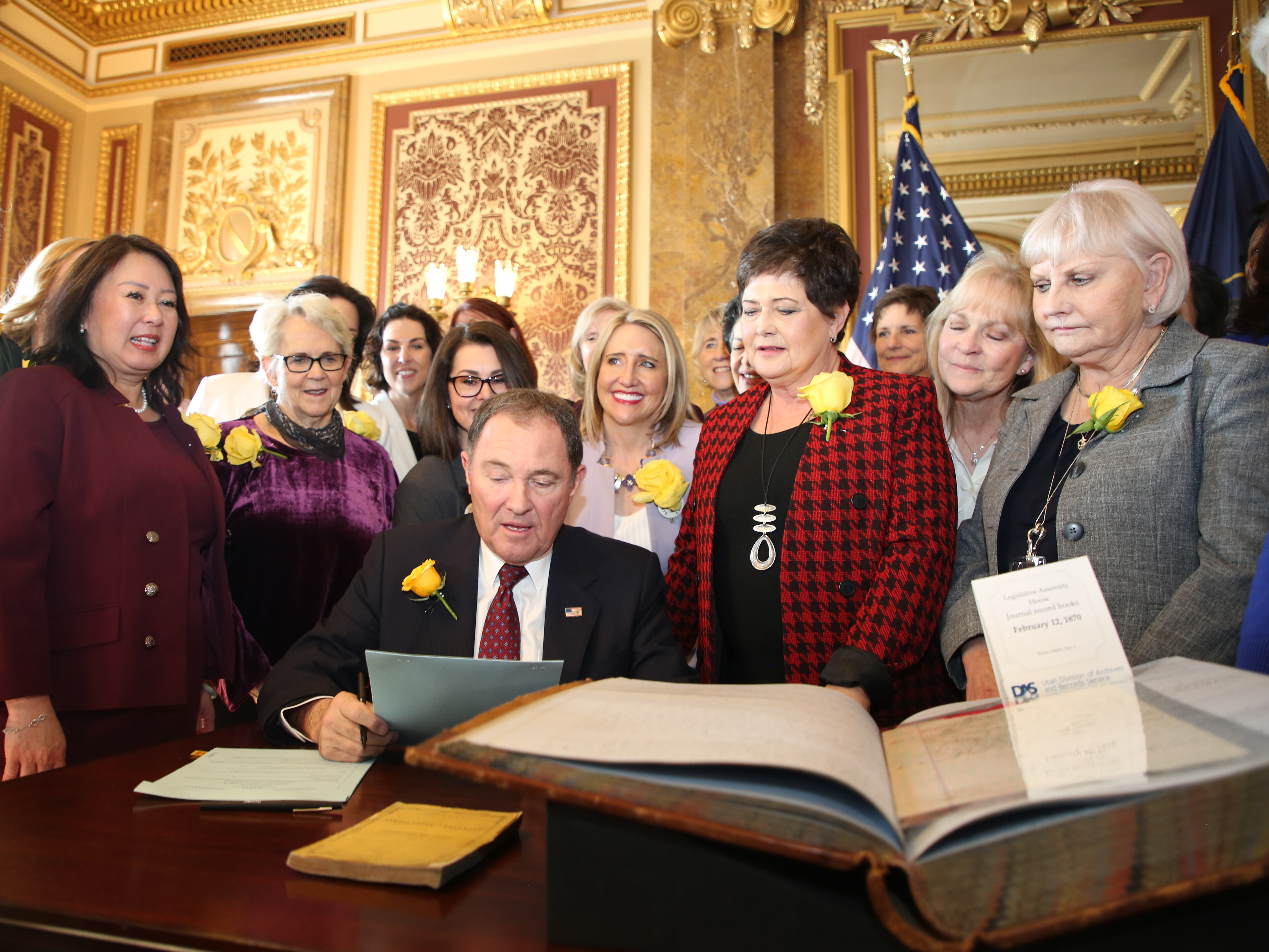 caption: Utah Gov. Gary Herbert signs bill honoring the state's pioneering women suffragists on Wednesday. He's surrounded by state senators and representatives, and his wife, who are all wearing the yellow rose symbolizing suffrage.<em> </em>