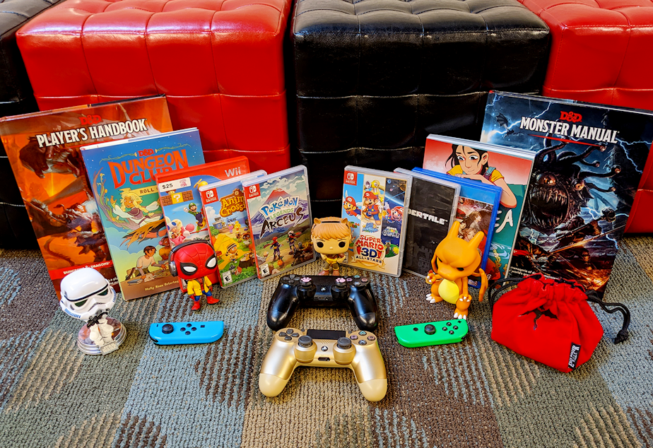 caption: Various pieces of pop culture media and memorabilia lie in front of red and black storage cubes. The assortment includes D&D rulebooks, graphic novels, video game disc cases, PS4 controllers, and Funko Pop figurines.