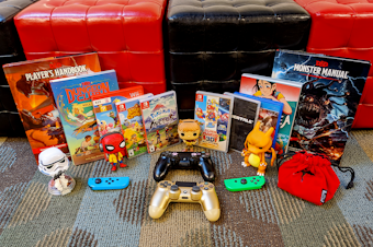 caption: Various pieces of pop culture media and memorabilia lie in front of red and black storage cubes. The assortment includes D&D rulebooks, graphic novels, video game disc cases, PS4 controllers, and Funko Pop figurines.