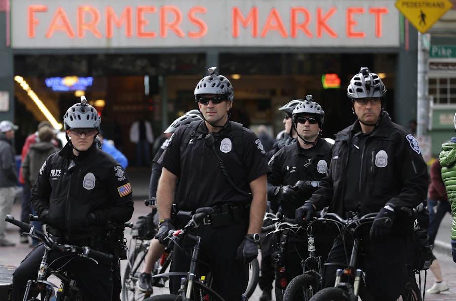 With a “FARMERS MARKET” sign in the background, a group of police officers in gray helmets and black uniforms stand on bikes. 