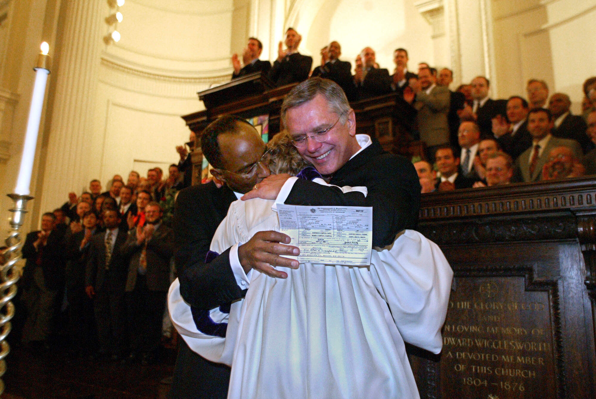 caption: David Wilson (left) and Robert Compton hug the Rev. Kim Crawford Harvie after she signed their marriage certificate following the ceremony at Arlington St. Church.  The Boston Gay Men's Chorus is in the background. (John Wilcox/MediaNews Group/Boston Herald via Getty Images)