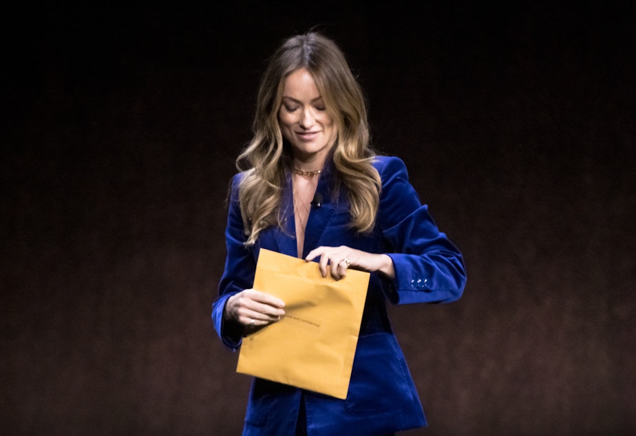 caption: Director and actress Olivia Wilde examines an envelope reading "Personal and Confidential" while onstage during the CinemaCon convention in Las Vegas on April 26. A man from the audience had slid the mysterious envelope toward her. It turns out she had been served with legal documents in an atypically public way.