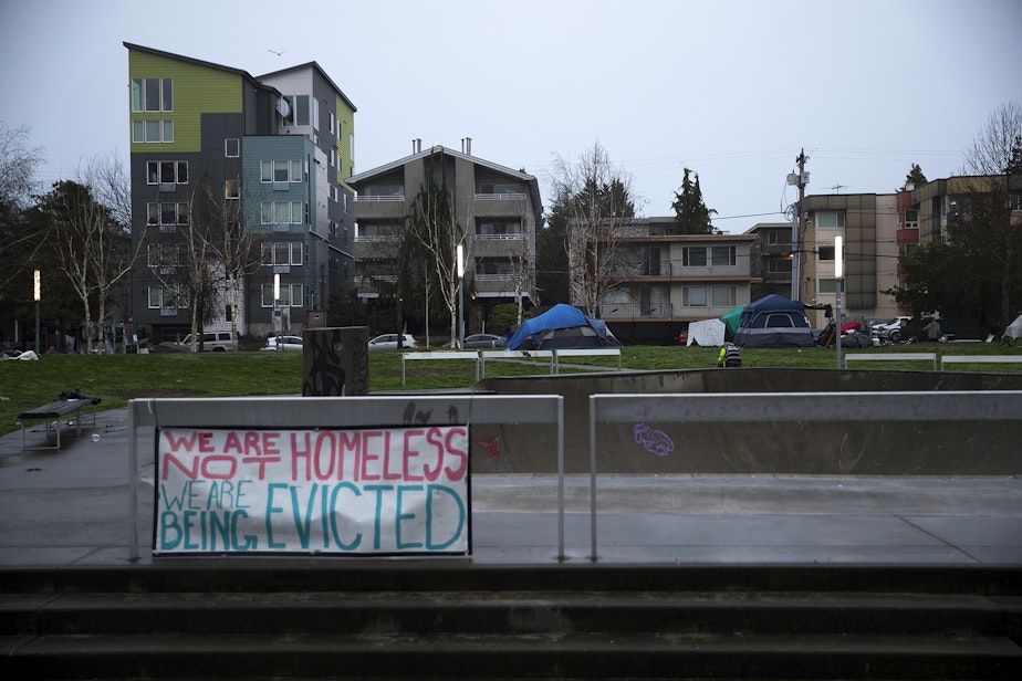 caption: 'We are not homeless, we are being evicted,' reads a sign in the foreground as Seattle Parks and Recreation employees prepare to sweep unhoused community members from Ballard Commons Park on Tuesday, December 7, 2021, in Seattle. 