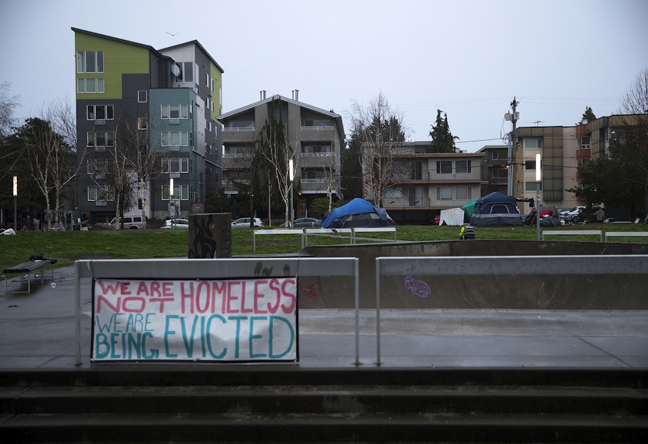 caption: 'We are not homeless, we are being evicted,' reads a sign in the foreground as Seattle Parks and Recreation employees prepare to sweep unhoused community members from Ballard Commons Park on Tuesday, December 7, 2021, in Seattle. 