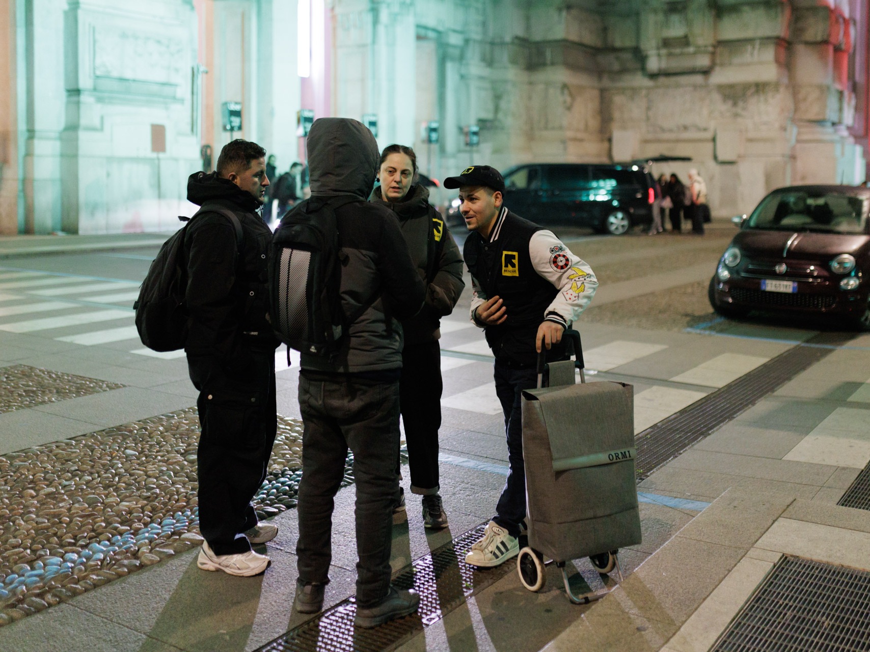 caption: Outreach team members of an International Rescue Committee (IRC), an international humanitarian group, stand outside Milano Centrale railway station at night. The station remains a critical hub for migrants and homeless individuals seeking temporary refuge from the winter cold.