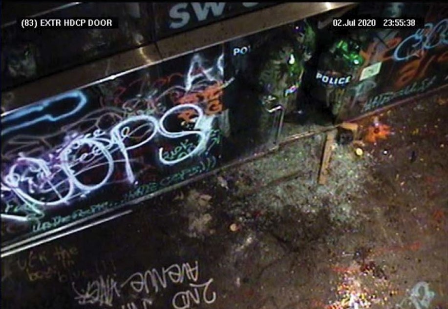 caption: In this July 2, 2020, photo released by the U.S. Attorney Office in Oregon and taken from security cameras, officers with the Federal Protective Service can be seen, top right, holding their shields up and guarding the doorway of the Hatfield Federal Courthouse after the glass door shattered.