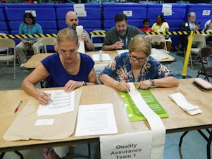 caption: Miami-Dade County Elections Department employees check the tabulation made by voting equipment that will be used in the upcoming state primary election.