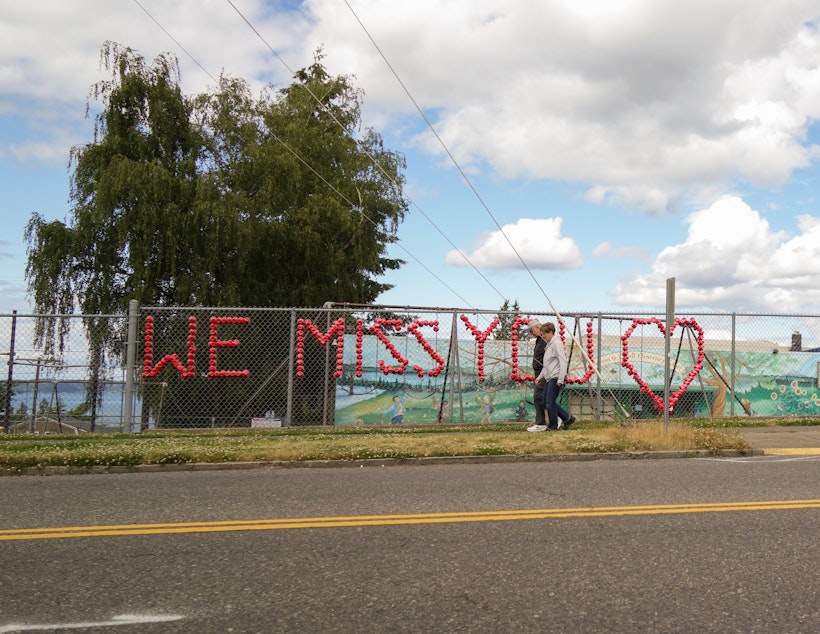 caption: A couple walks past the words "We Miss You" written with red plastic cups attached to the fence of Lowell Elementary School in Tacoma's North End. Tacoma Public Schools are preparing for students to return to in-person classes in the fall, but plans are also in place for remote learning in case of an outbreak at a school. In the meantime, the school district faces a $7 million budget shortfall and laid off or cut hours for hundreds of paraeducators earlier this year.