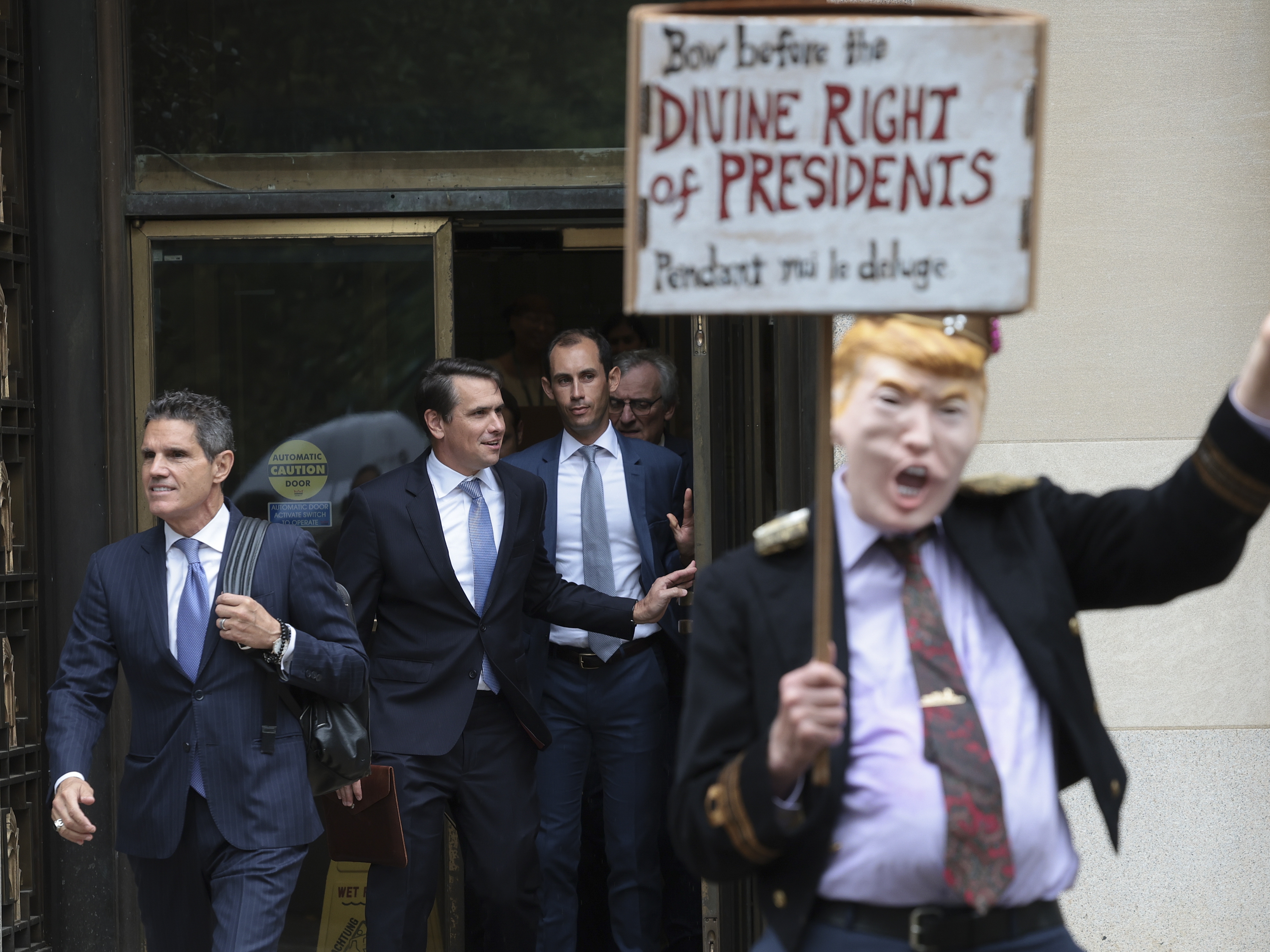 caption: Attorneys for former President Donald Trump, John Lauro (left) and Todd Blanche (second left), depart the E. Barrett Prettyman U.S. Courthouse in Washington, D.C., in 2023, near a protester wearing a costume in Trump's likeness.