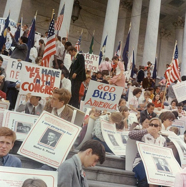 caption: Diane Benscoter, sitting with her head bowed beneath a "GOD BLESS AMERICA" sign, prays for then-President Richard Nixon not to be impeached. Benscoter was a member of the Unification Church, a religious, at the time.