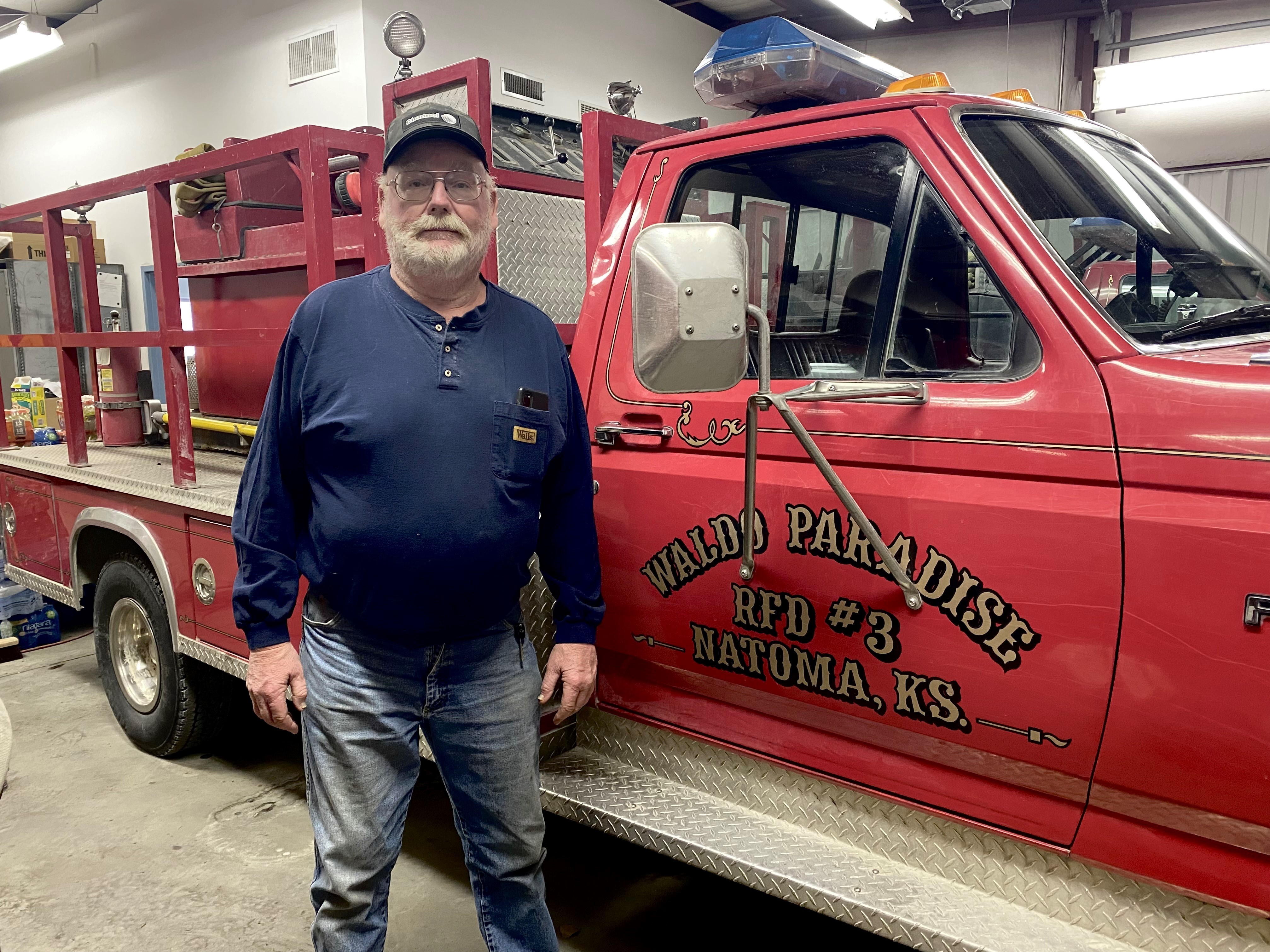 caption: Keith Koelling is 62 and still fighting fires. He says he worked 40 hours straight on a recent fire, only to find out he had COVID-19 at the time. He's standing next to a 26-year-old fire truck.